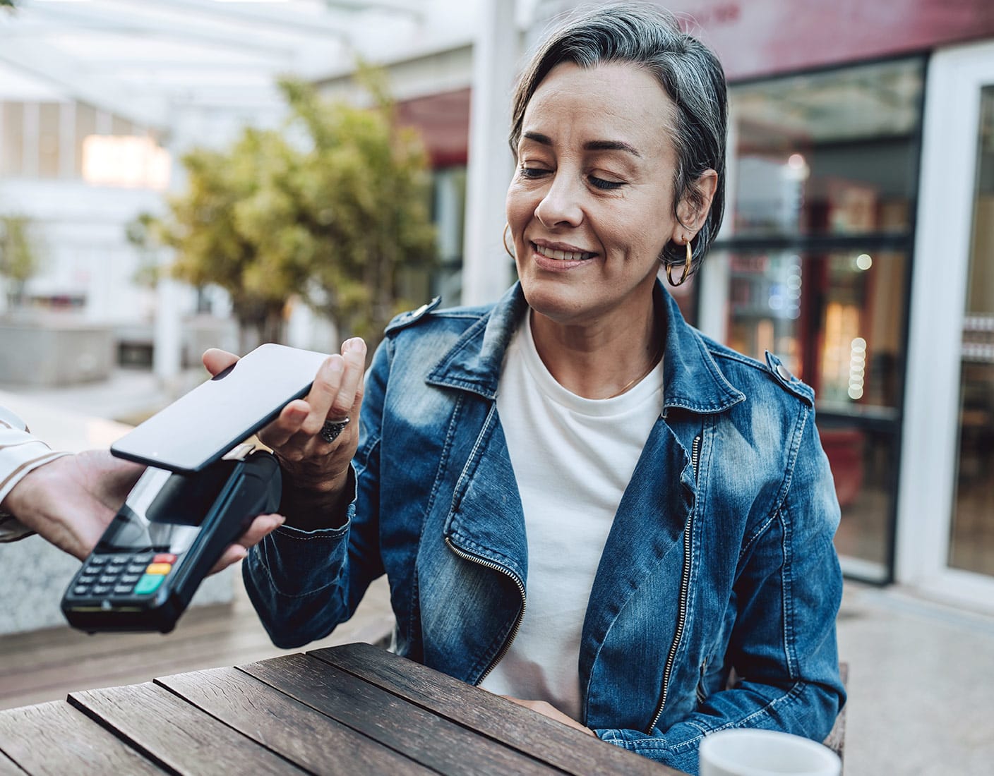 A smiling woman wearing denim jacket holds her phone over a tap and pay system.